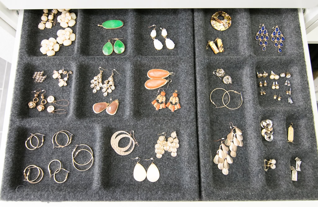 A drawer in an IKEA-inspired walk-in closet, lined with black fabric, displays an assortment of earrings. Various styles include hoops, studs, and dangling designs adorned with gemstones and metal accents in vibrant colors like green, orange, and blue, all neatly organized into compartments.