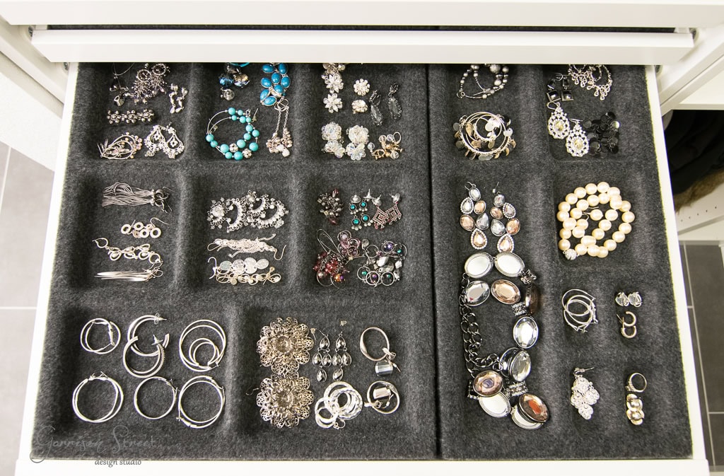 A neatly organized IKEA jewelry drawer displays various pieces, including rings, bracelets, earrings, and brooches on a dark, compartmentalized velvet liner. Nestled within a walk-in closet, the collection showcases a variety of colors and designs with intricate details and craftsmanship.