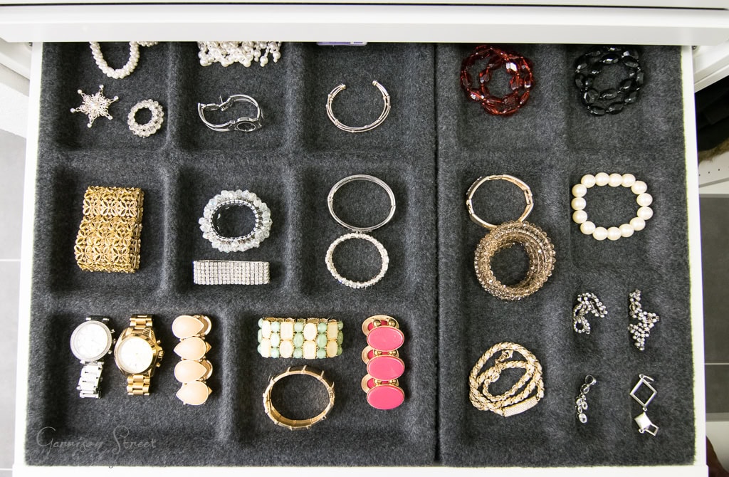 A neatly organized jewelry drawer inside the IKEA walk-in closet features various accessories, including bracelets, watches, earrings, and rings. Items are arranged in distinct sections on a soft, dark fabric for easy viewing and access.