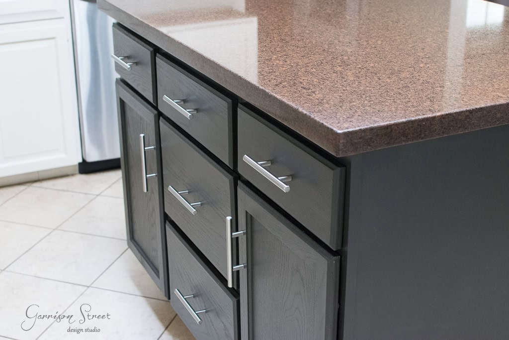 A modern kitchen island, crafted from base cabinets, boasts dark wood with sleek silver handles. Its countertop is a glossy, brown-speckled masterpiece, perfectly complementing the light beige tiled floor for a harmonious kitchen space.