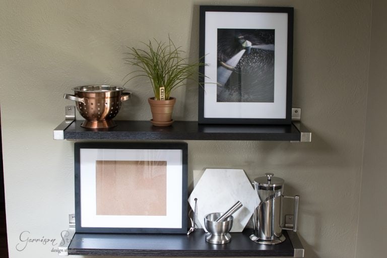 A wall shelf with two levels. The top shelf holds a copper colander, a potted plant, and a framed photo. The bottom shelf has a framed picture, a white hexagonal object, a French press, and a mortar and pestle.
