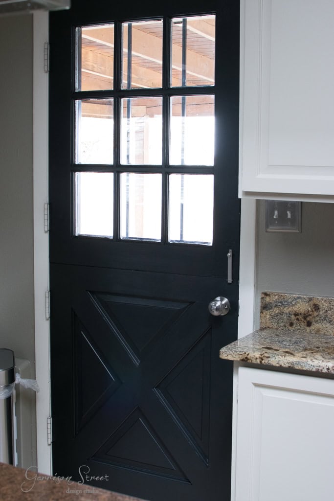 A black Dutch door with glass panels at the top half, allowing light to enter. The bottom half has a distinctive X design. A granite counter is visible on the right, and the door reflects a covered patio outside.