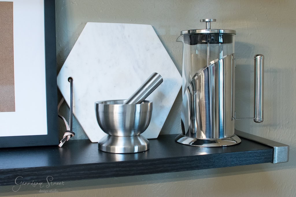 A stainless steel mortar and pestle sit beside a French press on a dark wooden shelf. Behind them, a white hexagonal marble trivet leans against the wall.