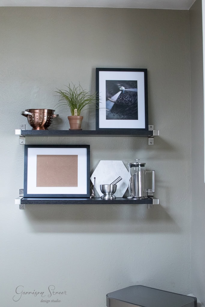 Two wall-mounted shelves with framed art, a potted plant, a copper pot, a marble cutting board, a metallic French press, and small kitchen items. The shelves are against a light gray wall, creating a minimalistic and modern look.