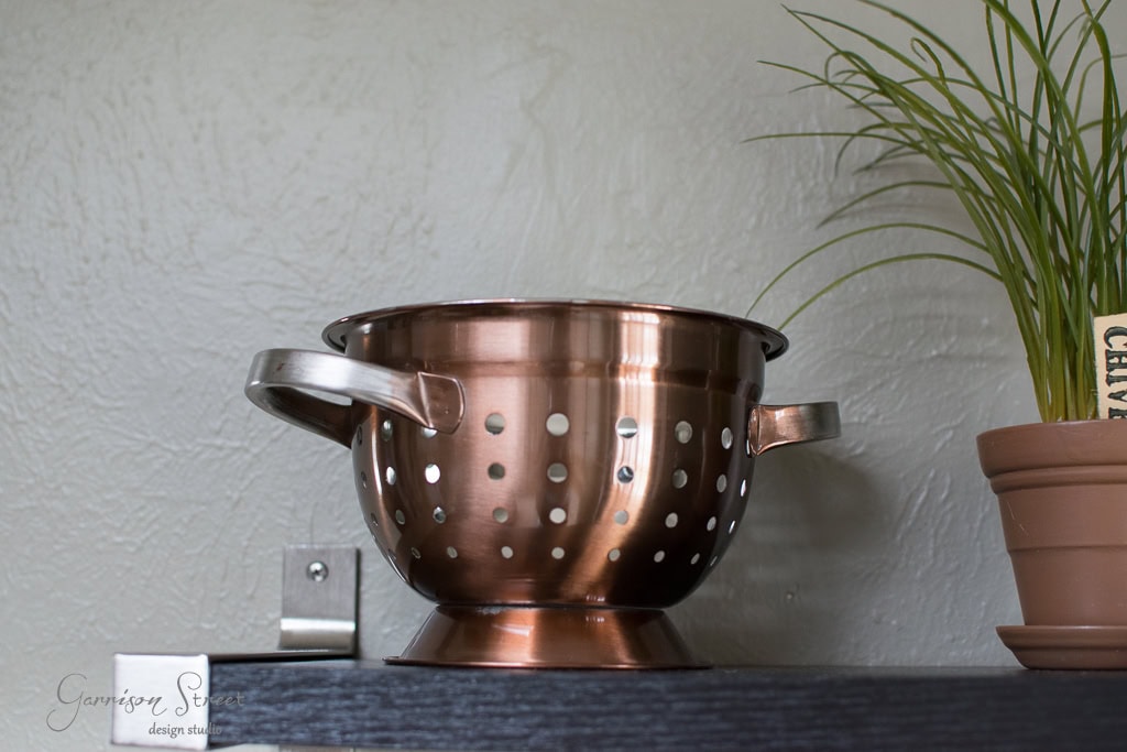 A copper colander with handles is placed on a dark wooden shelf. A potted plant with long, slender leaves is next to it, adding a touch of greenery to the scene. The wall behind is textured and light-colored.