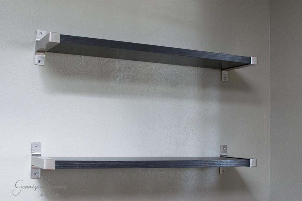 Two empty dark gray floating shelves mounted on a light-colored textured wall. The shelves have metal brackets for support, offering a minimalist and modern appearance.