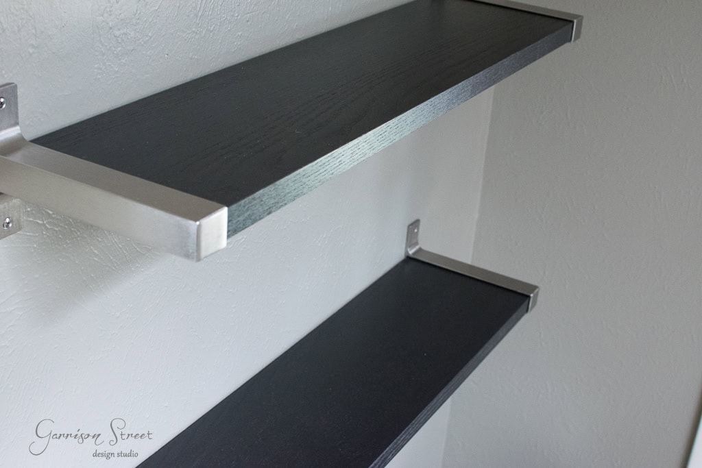 Close-up of two modern, dark wood floating shelves with metallic brackets on a light gray wall. The shelves are empty, showcasing their minimalist design.
