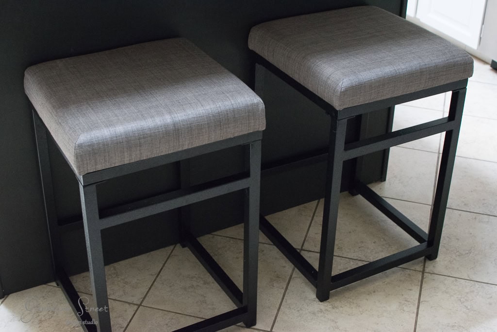 Two square bar stools with gray fabric cushions and black metal frames are positioned on a tiled floor beside a dark countertop.