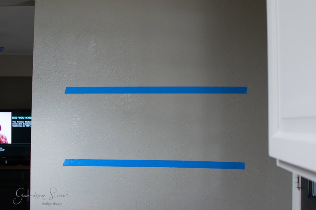 A beige wall with two parallel horizontal blue tape lines. A dark living room is partially visible on the left with a TV showing a person talking. A white door frame is on the right edge.