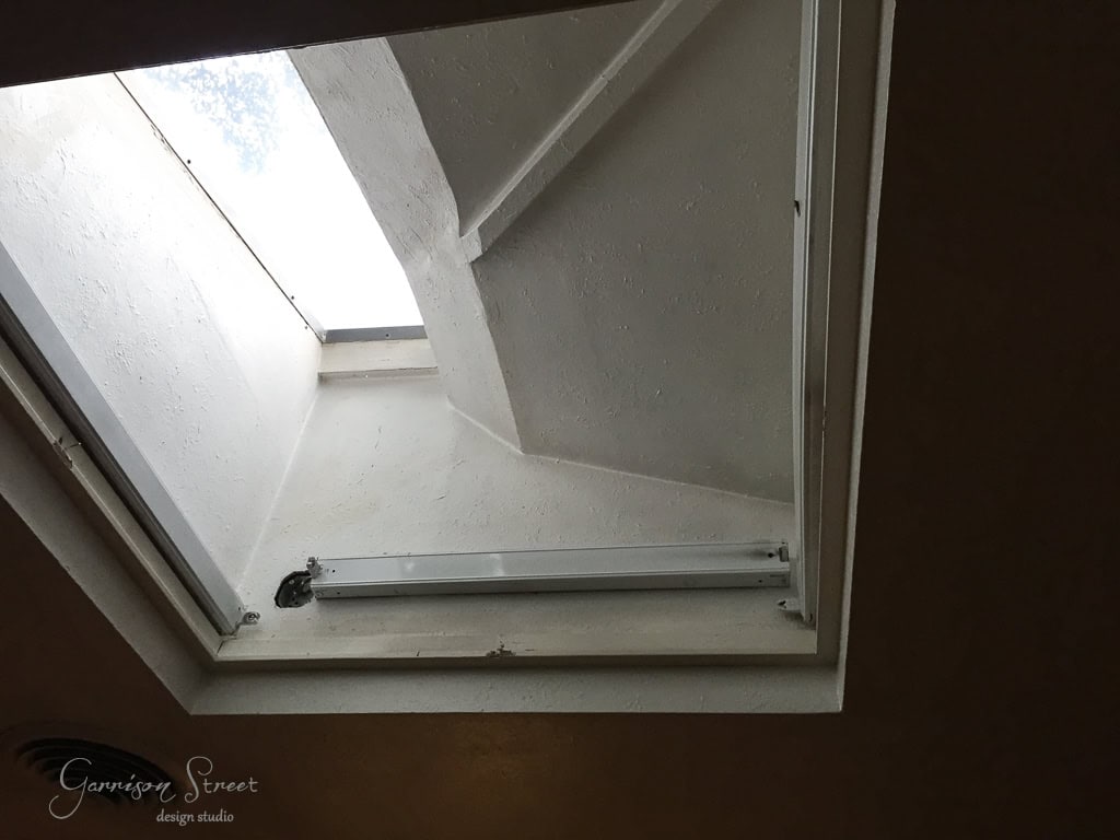A view from inside the kitchen looking up through an open skylight reveals the sky and a section of the roof. The skylight is framed by white walls, with a linear mechanism on the side for opening and closing, allowing natural light to flood the cooking space.