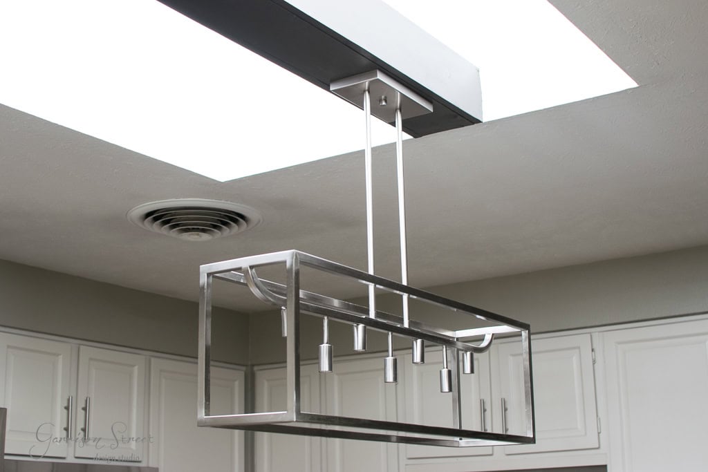 A modern rectangular metal chandelier with four light fixtures is suspended from a light gray ceiling featuring a recessed air vent. A skylight nearby illuminates white kitchen cabinets, enhancing the minimalist aesthetic.