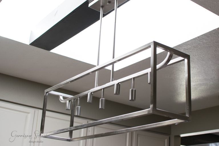 A modern rectangular metal chandelier hangs from the ceiling in the kitchen, beneath a skylight. The fixture features multiple cylindrical light sockets, enhancing the brightness of the room with its pristine white walls.
