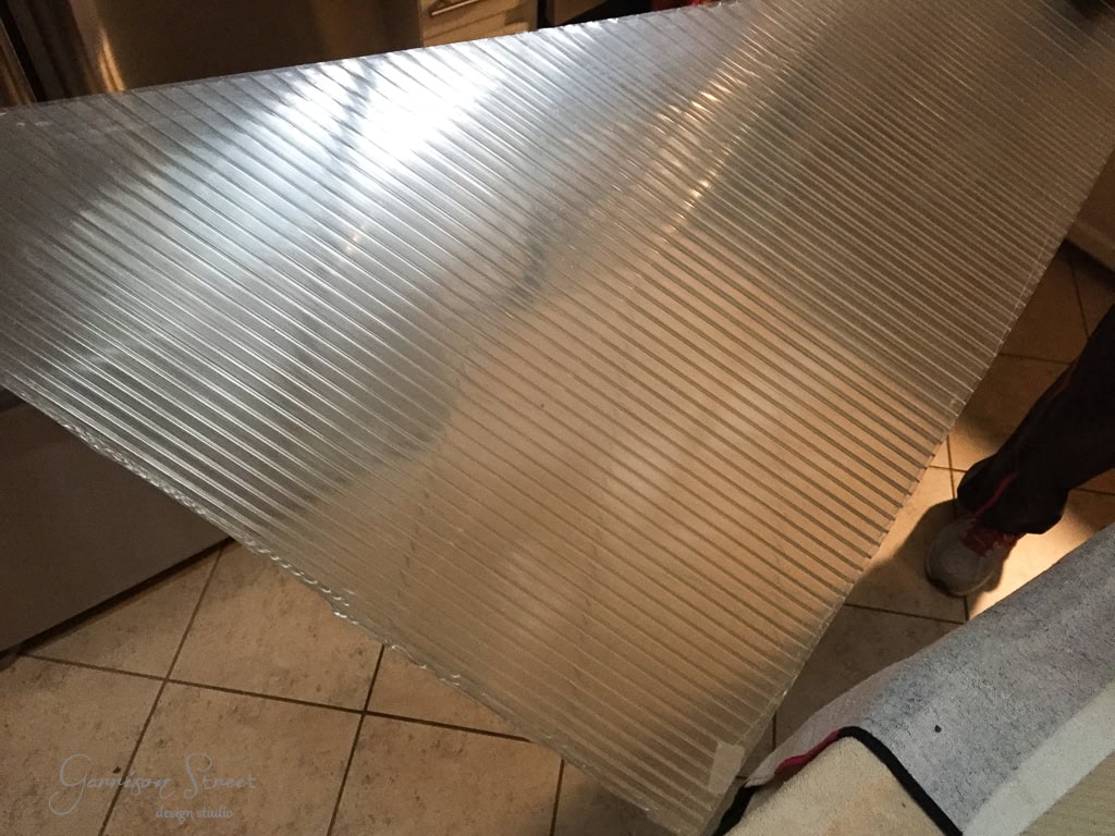 A person holds a large, translucent corrugated plastic sheet indoors beneath the kitchen skylight. The reflective, partially see-through sheet displays its corrugated ridges, while the beige-tiled floor gleams below. The person is partly visible, dressed in gray pants and shoes.