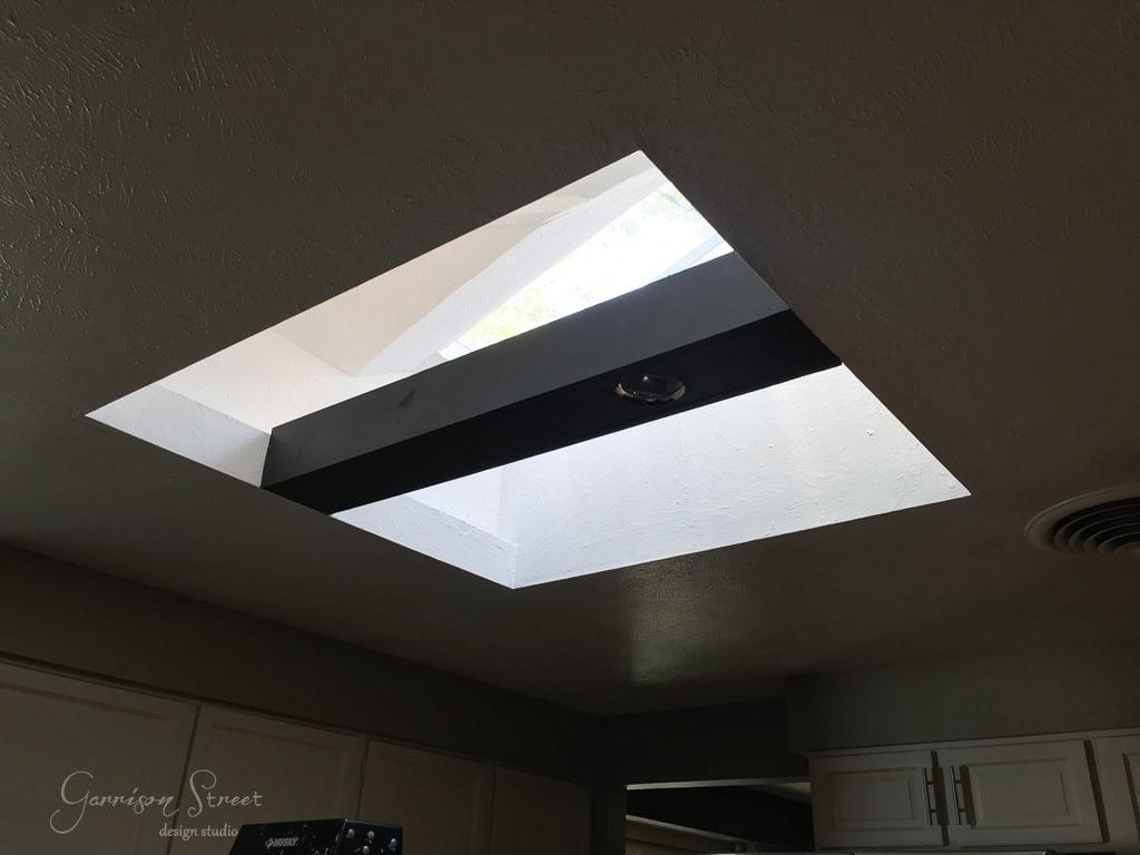 A ceiling with a large, square skylight beautifully illuminates the room with natural light, casting a striking contrast against the black beam that crosses it. The textured ceiling adds depth, while part of a sleek kitchen cabinet peeks out below.