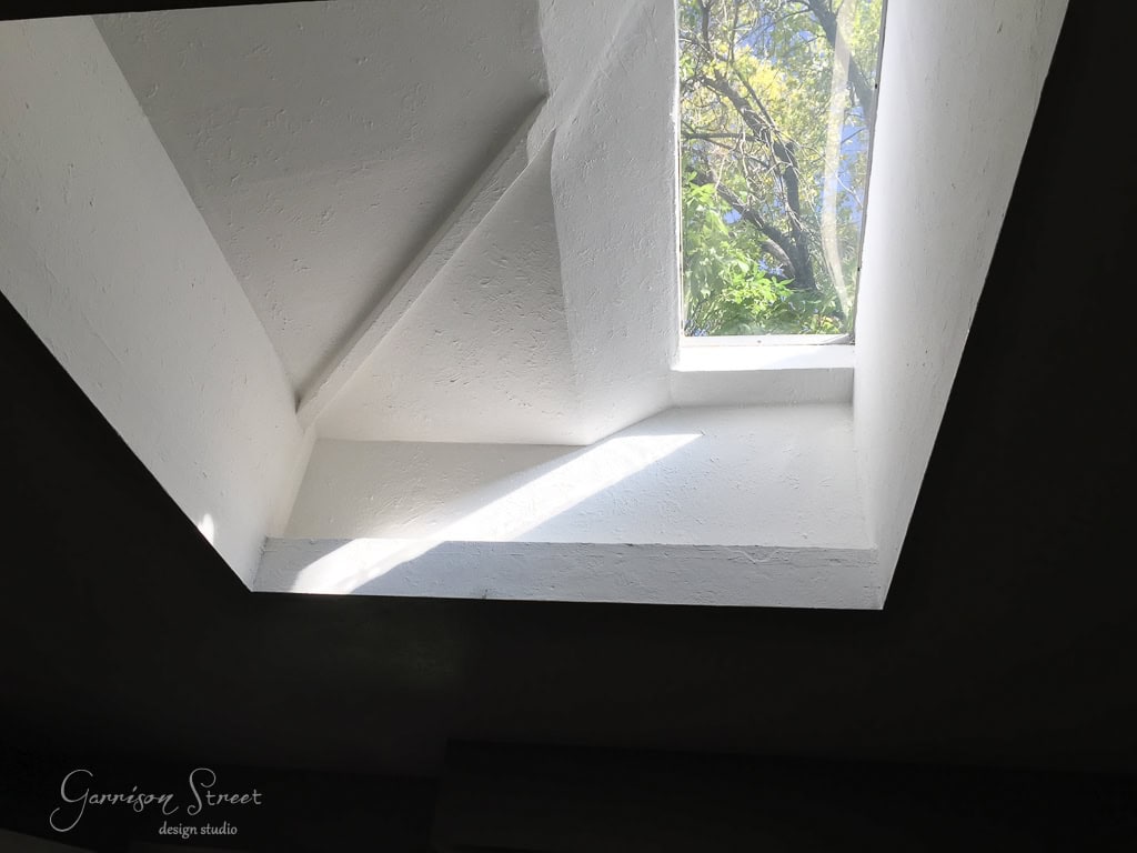 A skylight in the white ceiling of the kitchen allows sunlight to illuminate the interior space, with branches and green leaves visible through the window. Shadows create geometric shapes on the walls.