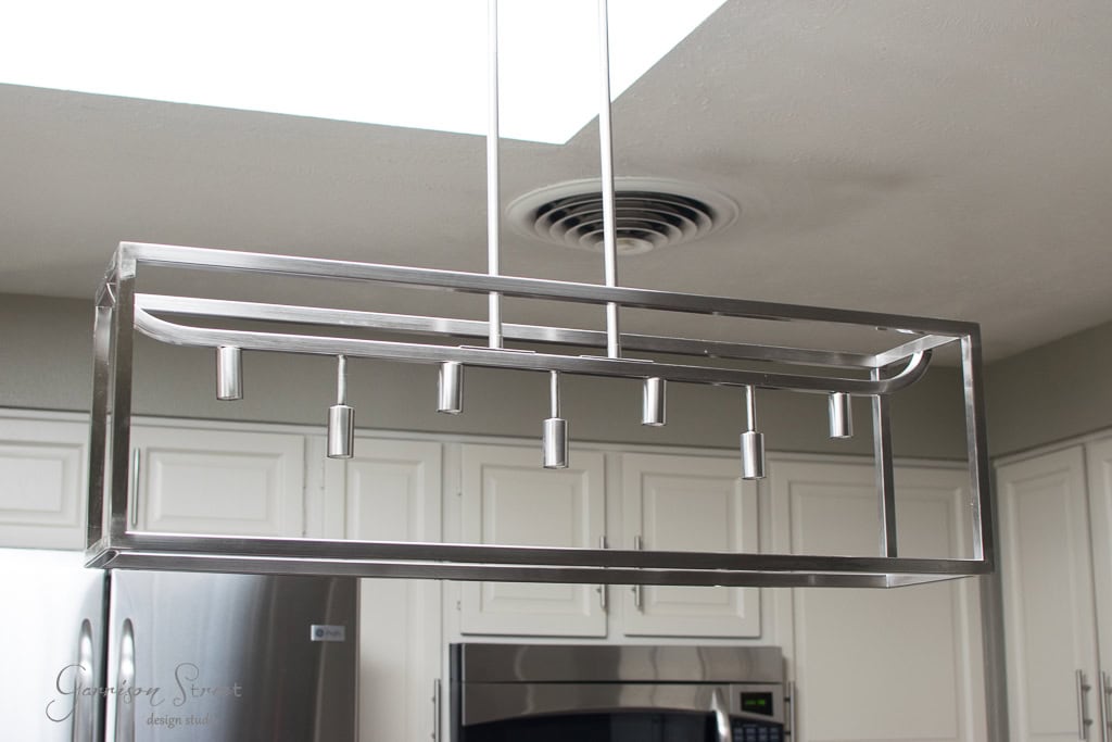 A modern rectangular metal light fixture with multiple bulb holders hangs from the ceiling in a kitchen, complemented by a skylight. White cabinets and a stainless steel refrigerator are visible in the background.