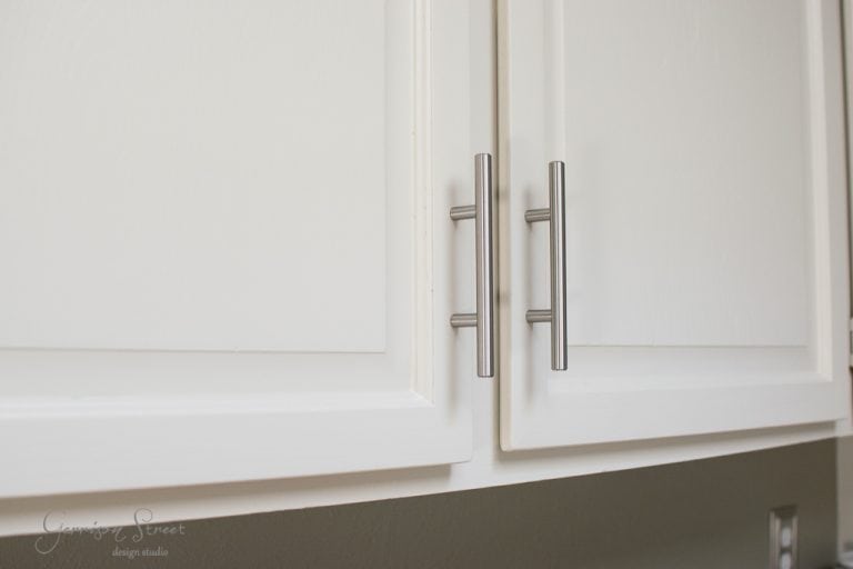 Close-up of white kitchen cabinet doors with long, silver vertical handles. The cabinets have a smooth, modern design.