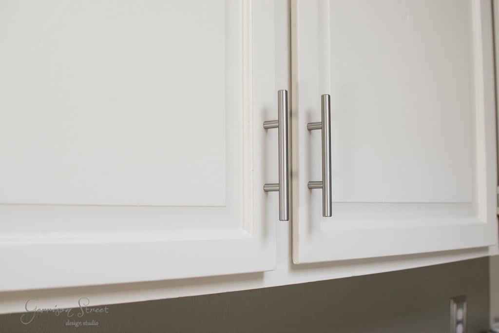 Close-up of white kitchen cabinet doors with long, silver vertical handles. The cabinets have a smooth, modern design.