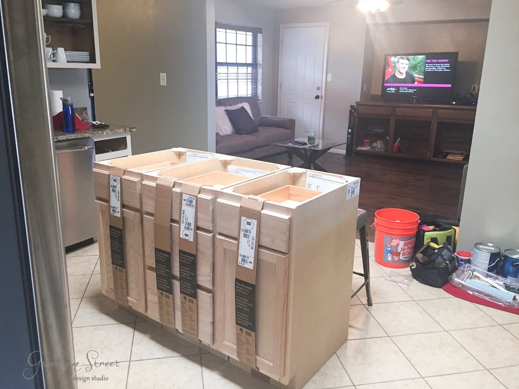 In the living room, a partially assembled wooden cabinet frame hints at a creative project in progress. With a sofa and TV in the background, tools, a bucket, and paint supplies scattered on the floor suggest tips on how to make a kitchen island out of base cabinets might be brewing.