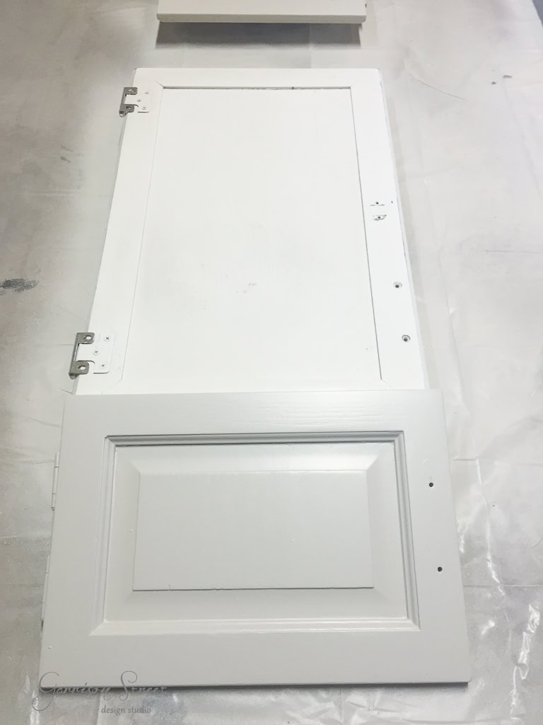 A white cabinet door lies flat on a surface, with visible hinges on the left. The door has a rectangular, beveled panel design. The surface is protected by a white covering.