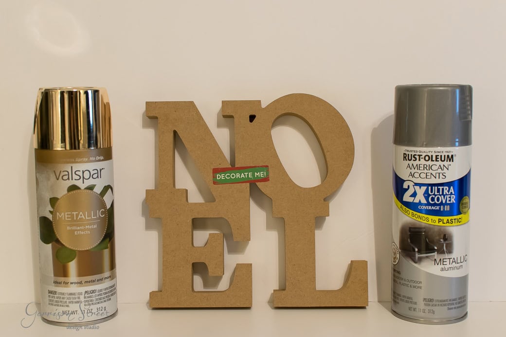 Two spray paint cans flank a wooden "NOEL" sign, perfect for your Christmas decor. On the left, a Valspar metallic gold can; on the right, a Rust-Oleum metallic silver can labeled for plastic. A small green label on the "NOEL" sign reads "DECORATE ME!.