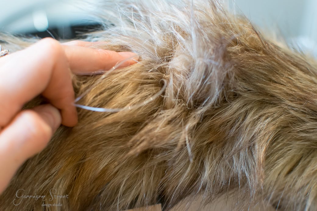 Close-up of a person handling soft, shaggy brown and white fur, reminiscent of a cozy Christmas stuffed animal. The fingers gently part the fur, revealing texture and layers in natural light, evoking warmth akin to a festive wreath.