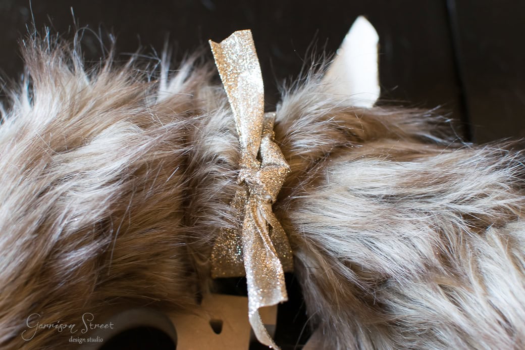 A close-up of a fuzzy faux fur object adorned with a shiny silver bow exudes festive charm, much like a Christmas wreath. The texture is soft and fluffy, with a hint of a white object subtly visible in the background.