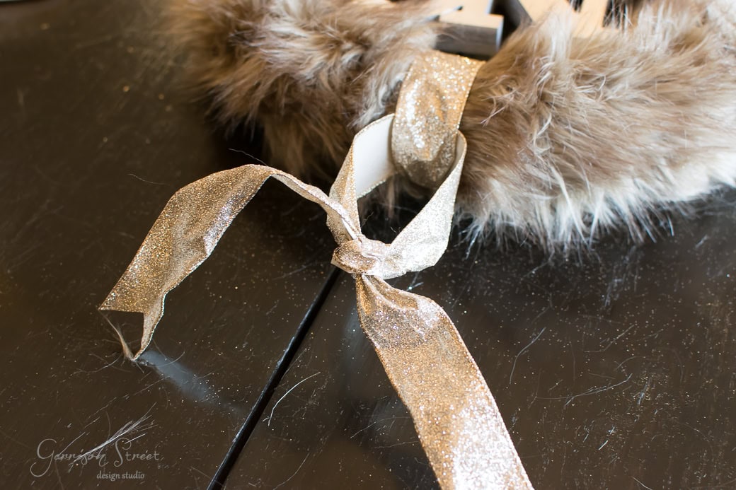 A close-up of a decorative golden ribbon tied in a bow, attached to a furry brown and white Christmas wreath on a reflective dark surface.