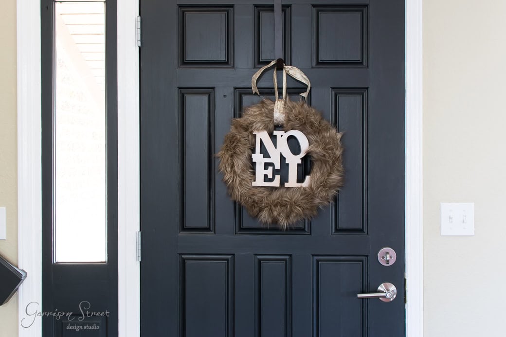 A black door with a round, fluffy fur wreath hanging on it. The festive wreath features the word "NOEL" in large white letters. The door is adorned with a silver doorknob and set against a light-colored wall, evoking a cozy Christmas atmosphere.