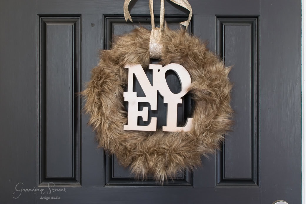 A dark door adorned with a fluffy, brown fur wreath. Wooden letters spelling "NOEL" are placed in the center of this Christmas decoration, hanging by a gold ribbon.