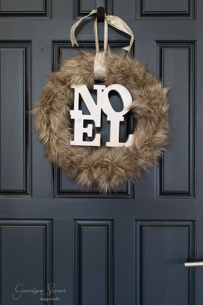 A dark wooden door adorned with a Christmas wreath resembling fluffy fur. The wreath prominently displays "NOEL" in large white letters, elegantly hung with a silver ribbon.