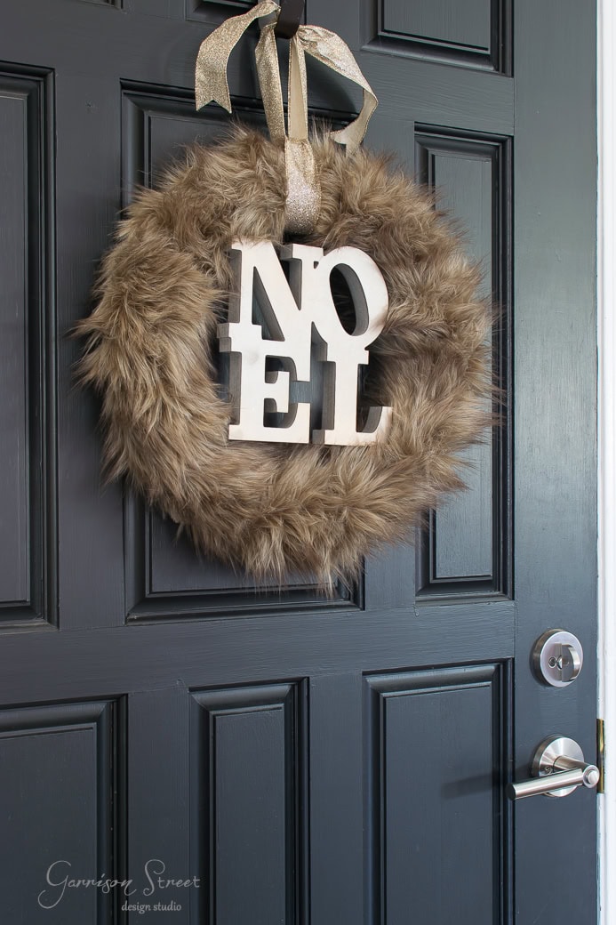 A dark-colored door adorned with a Christmas wreath, wrapped in ribbon and large white letters spelling "NOEL." The fluffy wreath adds a festive touch to the entrance.