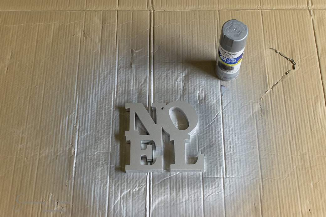 A cardboard surface with a spray-painted grey "NOEL" sign and a can of gray spray paint beside it. The cardboard shows a lighter area where the paint was applied, evoking the subtle charm of a minimalist Christmas wreath.