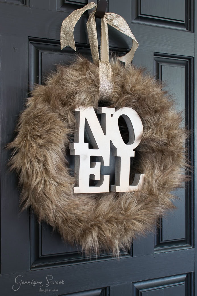 A Christmas wreath covered in brown fur hangs elegantly on a dark blue door. Adorned with a large "NOEL" sign in white letters, it's beautifully suspended by a sparkly gold ribbon.