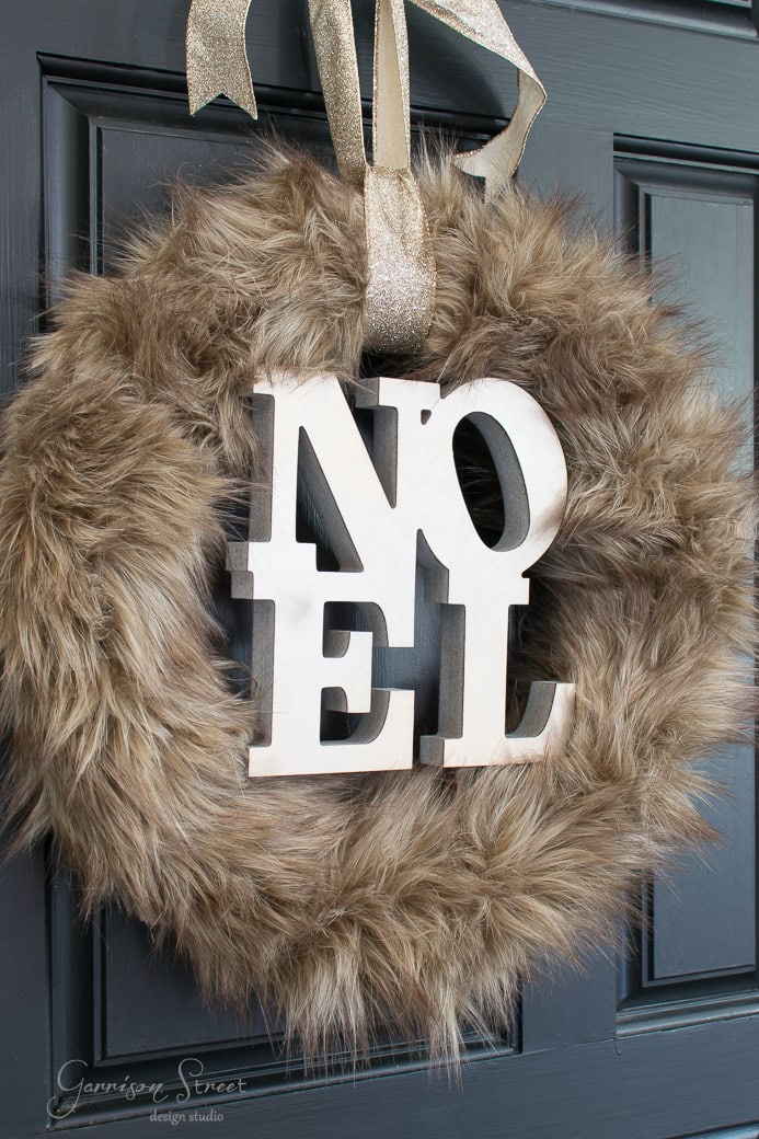 A fluffy, brown Christmas wreath adorned with large, white letters spelling "NOEL" hangs on a dark blue door. A shiny, gold ribbon is looped through the top of the wreath for hanging.