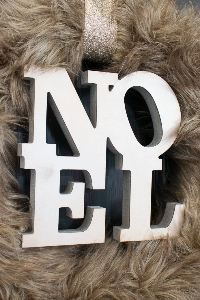 A wooden "NOEL" sign with a fur-like Christmas wreath around it, elegantly hanging from a glittery ribbon.