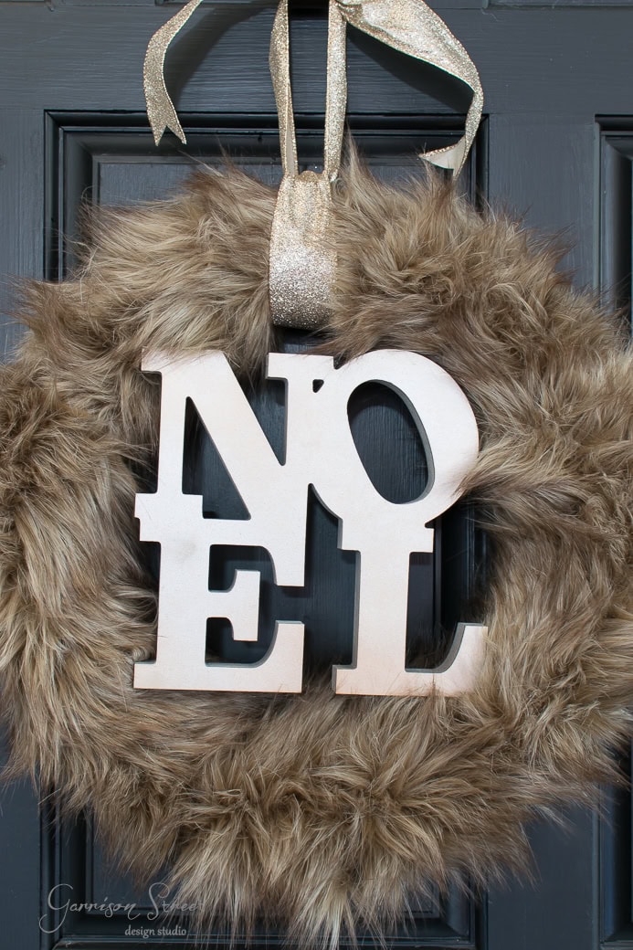 A decorative Christmas wreath made of faux fur with large wooden letters spelling "NOEL" hangs on a door. The festive wreath is adorned with a shimmering ribbon for hanging.