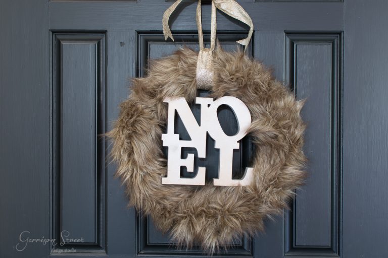A festive brown fur wreath with the word "NOEL" in white letters graces a dark blue door, bringing a touch of Christmas cheer. A shimmering gold ribbon loops through the top for hanging.
