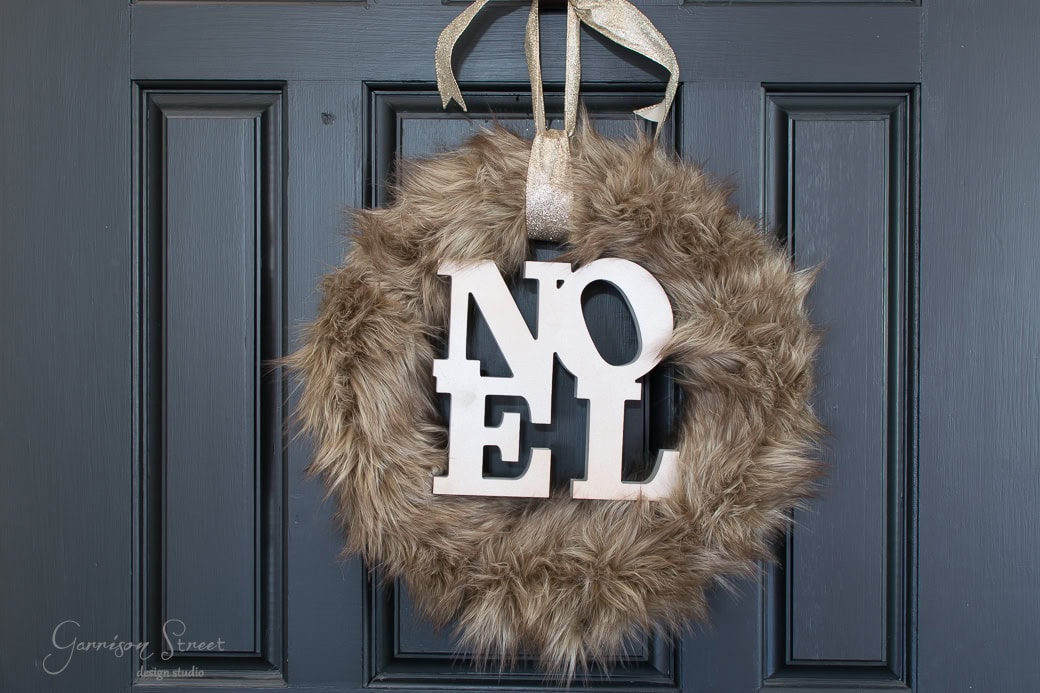 A festive brown fur wreath with the word "NOEL" in white letters graces a dark blue door, bringing a touch of Christmas cheer. A shimmering gold ribbon loops through the top for hanging.