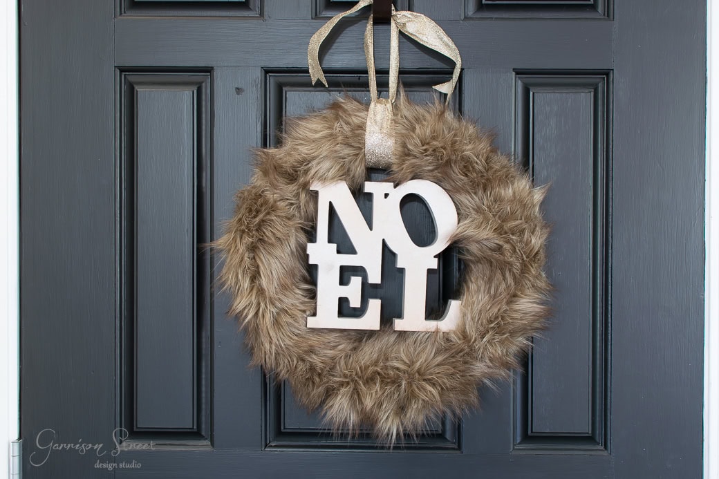 A festive Christmas wreath with the word "Noel" in large, white letters hangs on a dark-colored door. The wreath is adorned with soft, brown faux fur and is suspended by a gold ribbon.