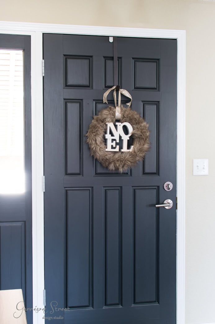 A black front door adorned with a festive Christmas wreath, crafted from brown fur and hanging elegantly from a ribbon. White letters spell "NOEL," illuminated gently by the light streaming through a nearby window.