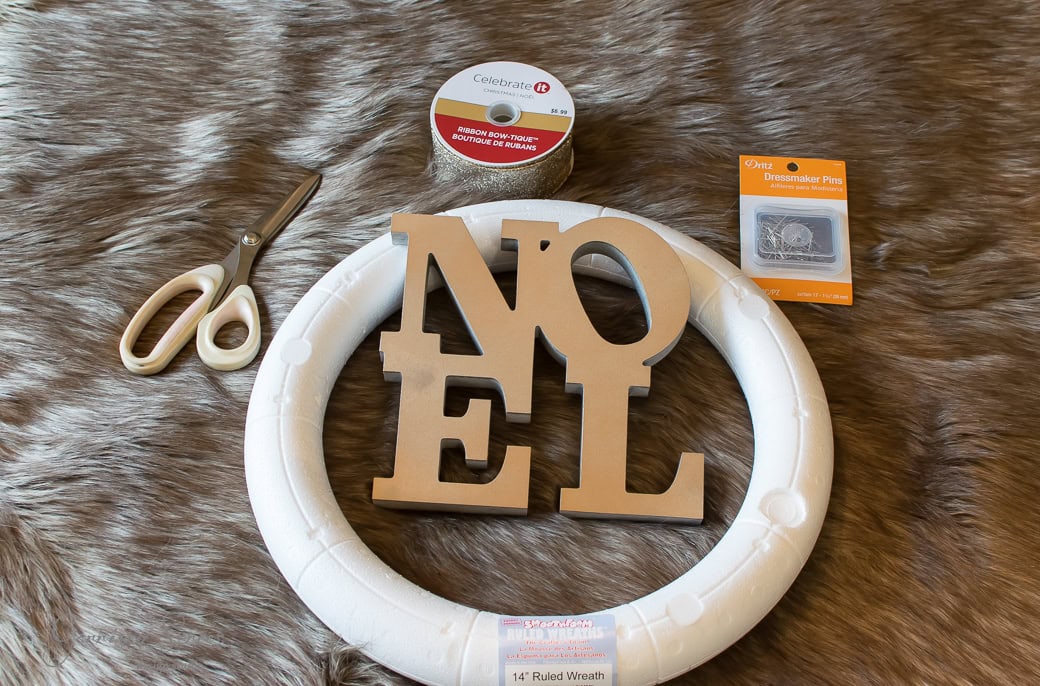 Crafting materials on a furry surface: scissors, a roll of festive red ribbon, wooden "NOEL" letters, and a white foam wreath base ready for Christmas magic. Complete with dressmaker pins, this scene invites the creation of holiday cheer.