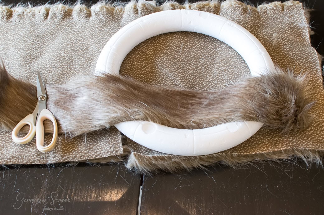 A crafting project in progress: a white foam ring gradually transforming into a cozy Christmas wreath, partially wrapped with brown faux fur. Scissors are poised on the furry fabric beside the ring, resting on a black table.