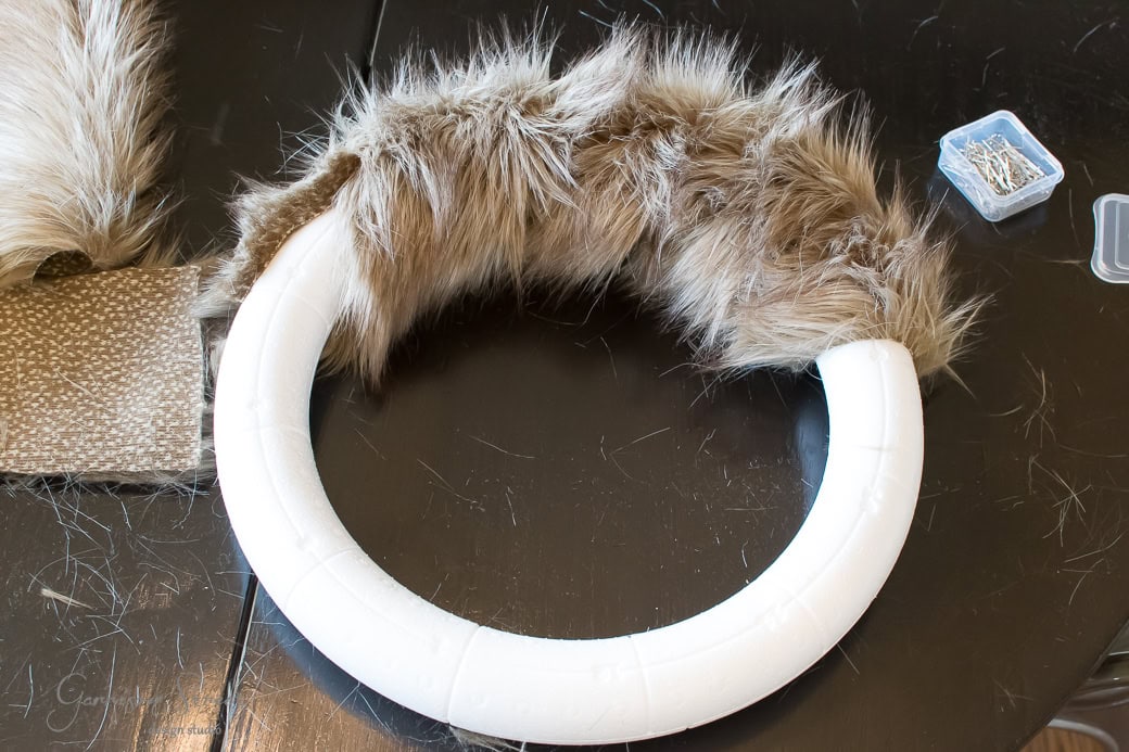 A crafting project on a black table features a wreath in progress, with a white foam ring partially wrapped in brown faux fur. Nearby, a piece of fabric and a small box of pins suggest preparations for a cozy Christmas decoration.