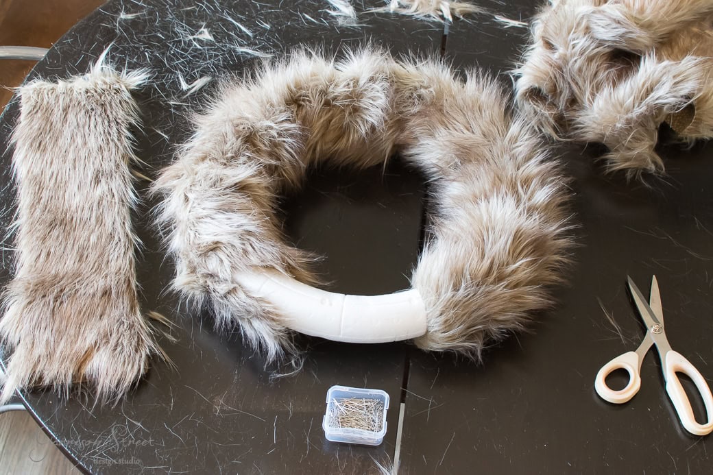 A festive crafting scene unfolds on the table: a wrapped circular base resembling a Christmas wreath, adorned with brown and beige faux fur. Nearby lie faux fur fabric pieces, scattered straight pins, and a trusty pair of scissors.