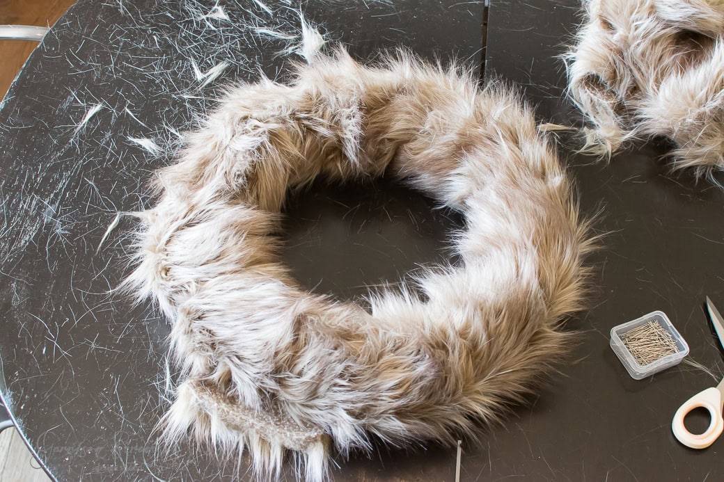 A fluffy, circular Christmas wreath made of soft, light brown faux fur is laid on a scratched black table. Nearby, there is a container of pins and a pair of scissors.
