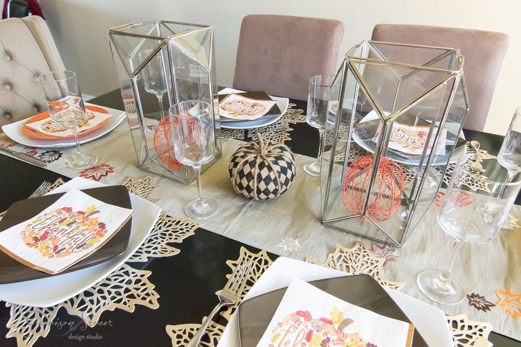 A dining table set for fall, featuring geometric lanterns, woven pumpkins, and Thanksgiving-themed plates on a decorative runner with intricate leaf patterns that perfectly capture the Thanksgiving spirit.