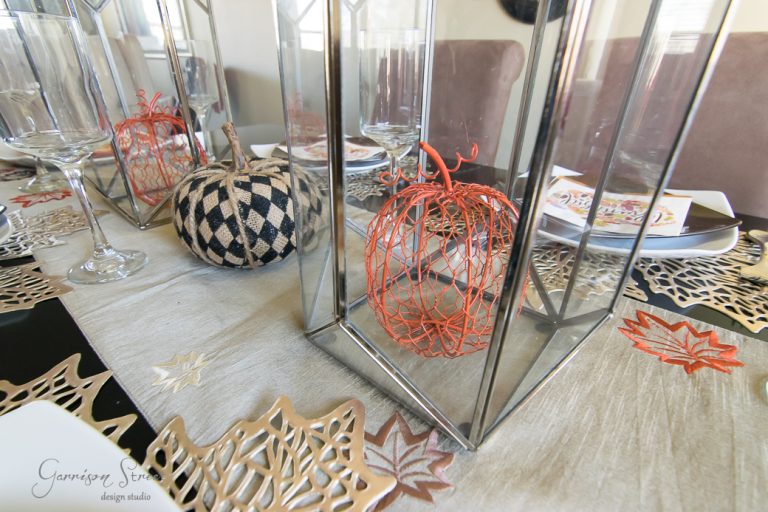 A table is elegantly decorated for Thanksgiving, featuring a patterned black and white pumpkin and a wire orange pumpkin nestled inside lanterns. The tablecloth boasts fall leaf designs in white, beige, and rust. Wine glasses and plates are neatly arranged beside the lanterns.