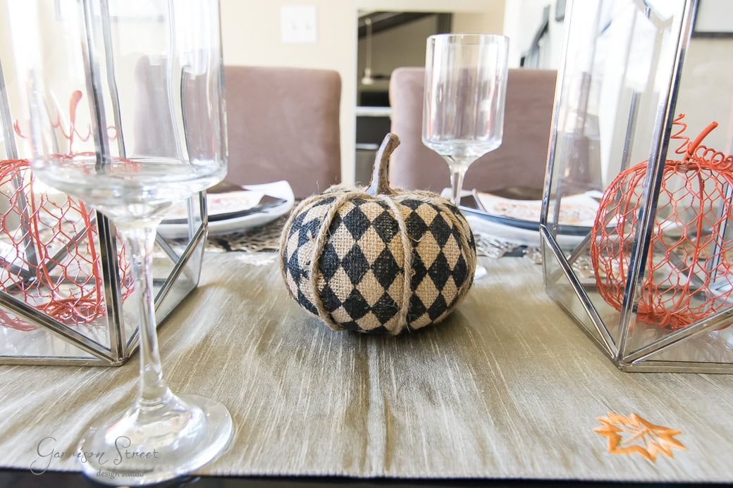 A dining table set for fall, perfect for Thanksgiving, features a centerpiece with a black and white checkered pumpkin nestled between glass holders containing orange woven ornaments. Wine glasses and plates are neatly arranged to welcome guests.