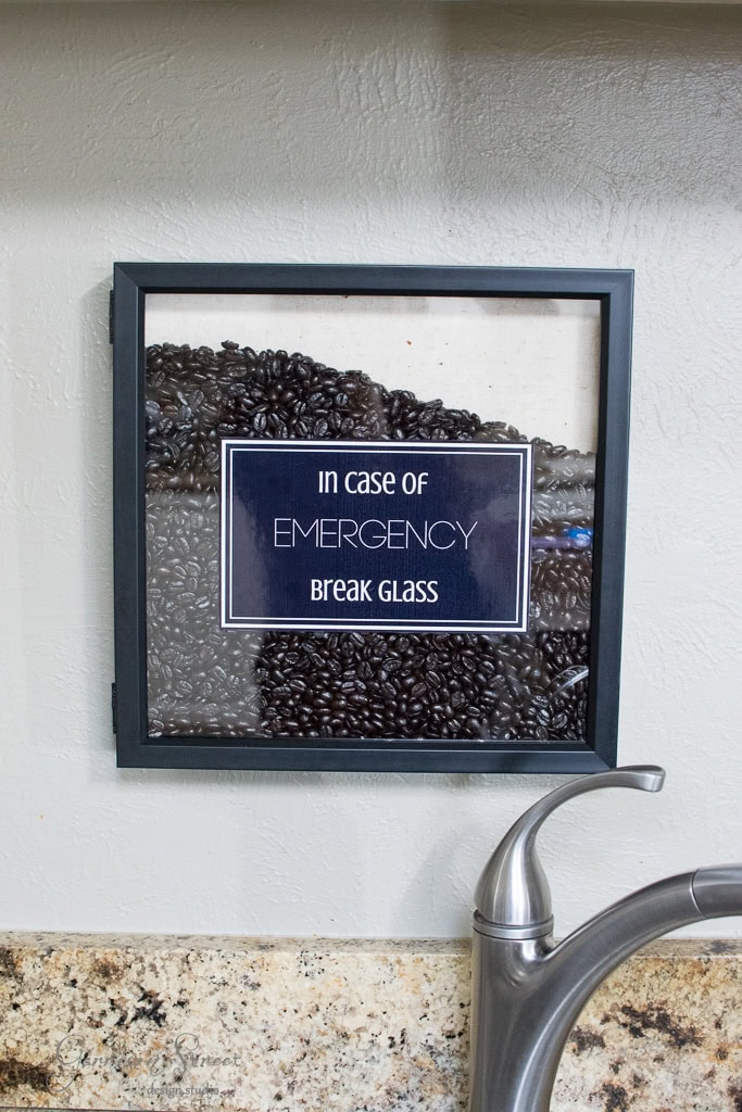 A framed glass box filled with coffee beans hangs on a wall above a kitchen counter. The sign inside reads "In Case of Emergency Break Glass." A stainless steel faucet is visible in the foreground.